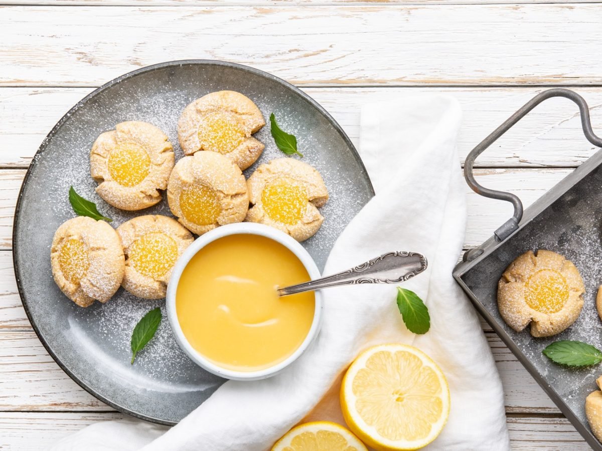 Die Lemon Curd Plätzchen auf einem grauen Teller und weißem Holztisch. Drumherum Zitronen und Blätter der Pflanze zusammen mit den Grundzutaten als Deko. Alles direkt von oben fotografiert.