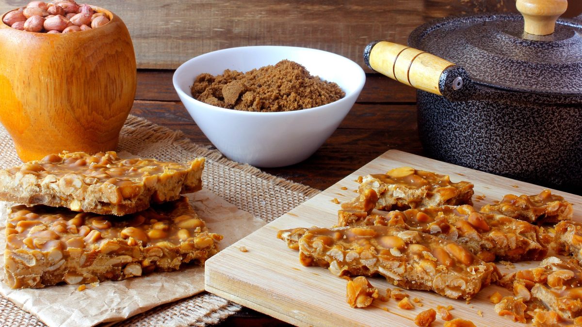 Pé-de-Moleque-Stücke auf Holzbrettern, daneben ein Steinguttopf und zwei Schalen mit braunem Zucker und Erdnüssen.