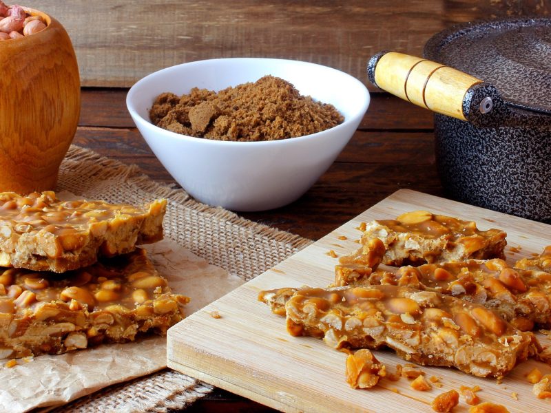 Pé-de-Moleque-Stücke auf Holzbrettern, daneben ein Steinguttopf und zwei Schalen mit braunem Zucker und Erdnüssen.
