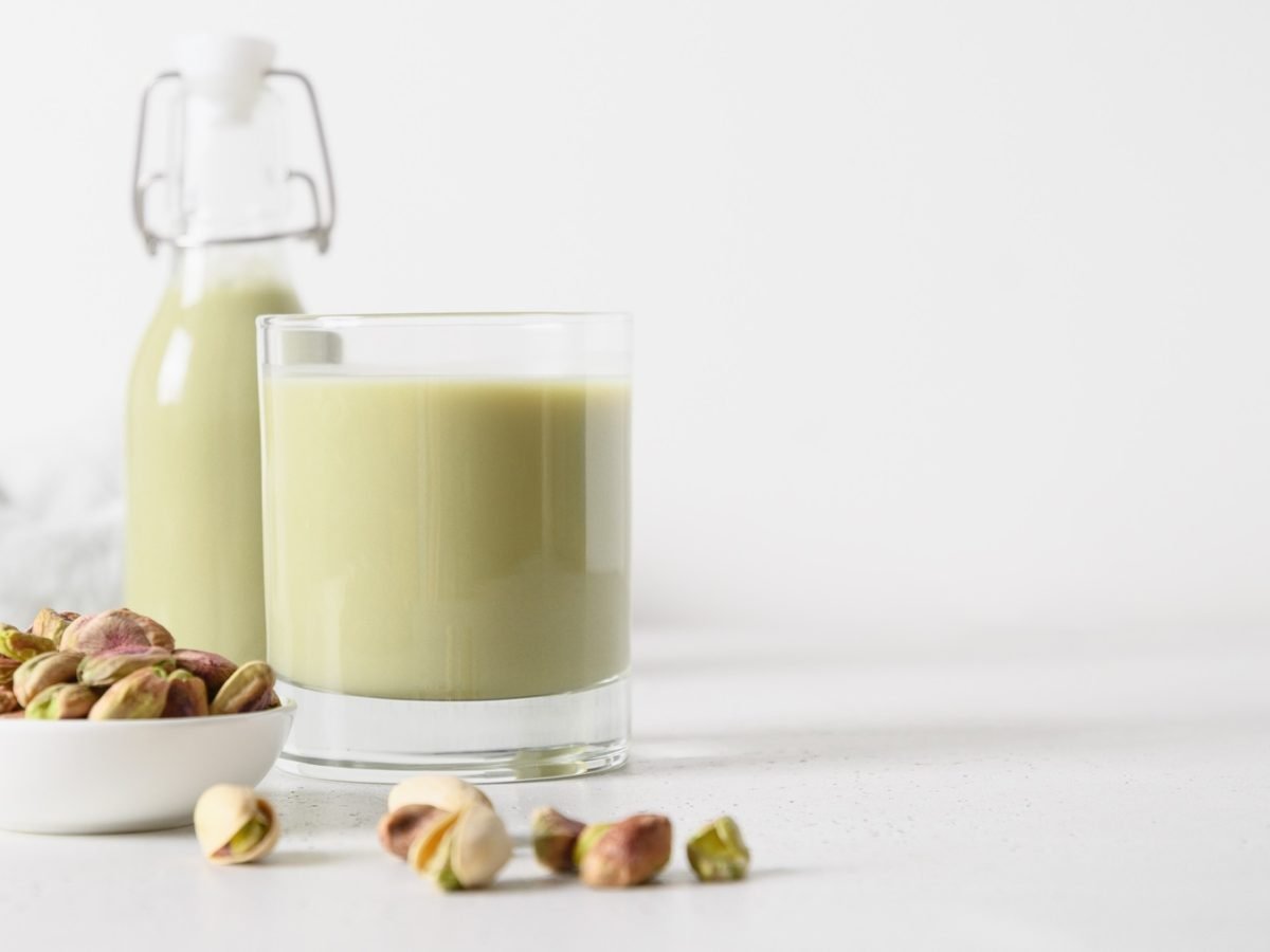 Ein Glas und eine Flasche mit Pistazienmilch vor einem weißen Hintergrund, im Vordergrund frische Pistazien.