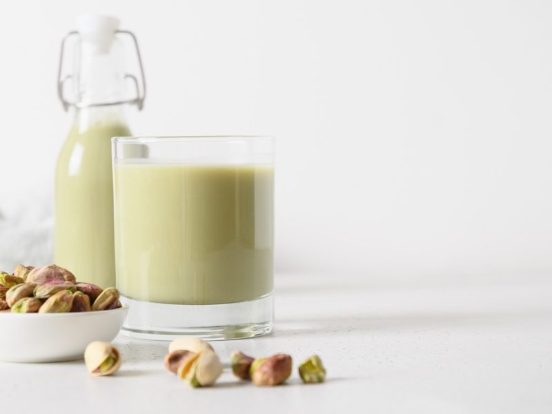Ein Glas und eine Flasche mit Pistazienmilch vor einem weißen Hintergrund, im Vordergrund frische Pistazien.