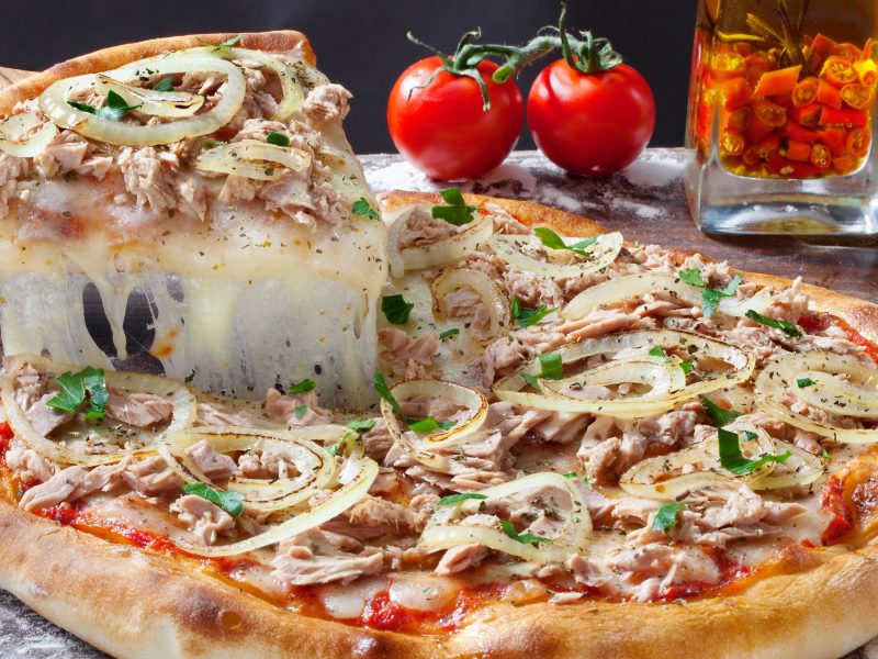 Eine Pizza Tonno e Cipolla, die auf einem Holztisch liegt, wird angeschnitten. Das Stück, dass angeschnitten wird, zieht sehr viel Käse. Im Hintergrund liegen frische Tomaten.