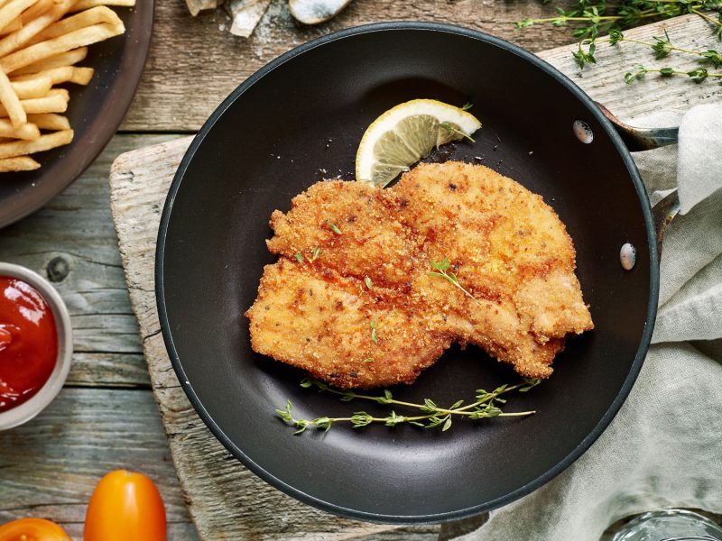 Thymian-Schnitzel auf schwarzem Teller. Daneben Zitrone und frischer Thymian, im Szenario Pommes und Ketchup.