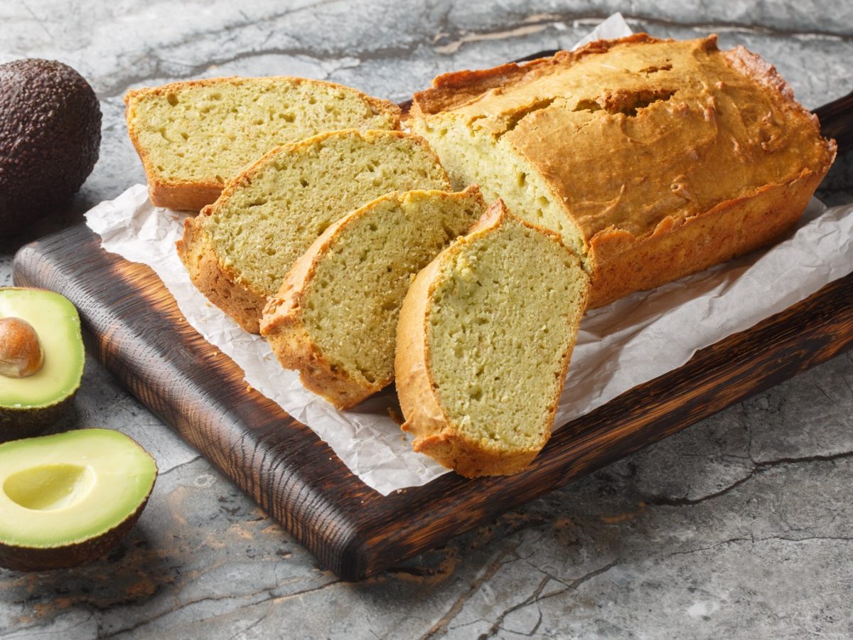 Angeschnittenes Avocadobrot auf einem Holzbrett, daneben eine halbierte Avocado und eine Ganze.