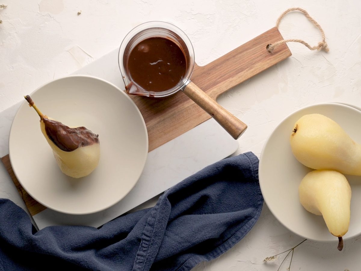 Ein weißer Teller mit der Birne und Schokosauce auf einem dunklen Brett und hellem Untergrund. Daneben auch noch ein kleiner Topf mit Schokosauce und ein weiterer Teller mit zwei pochierten Birnen. Alles direkt von oben fotografiert.
