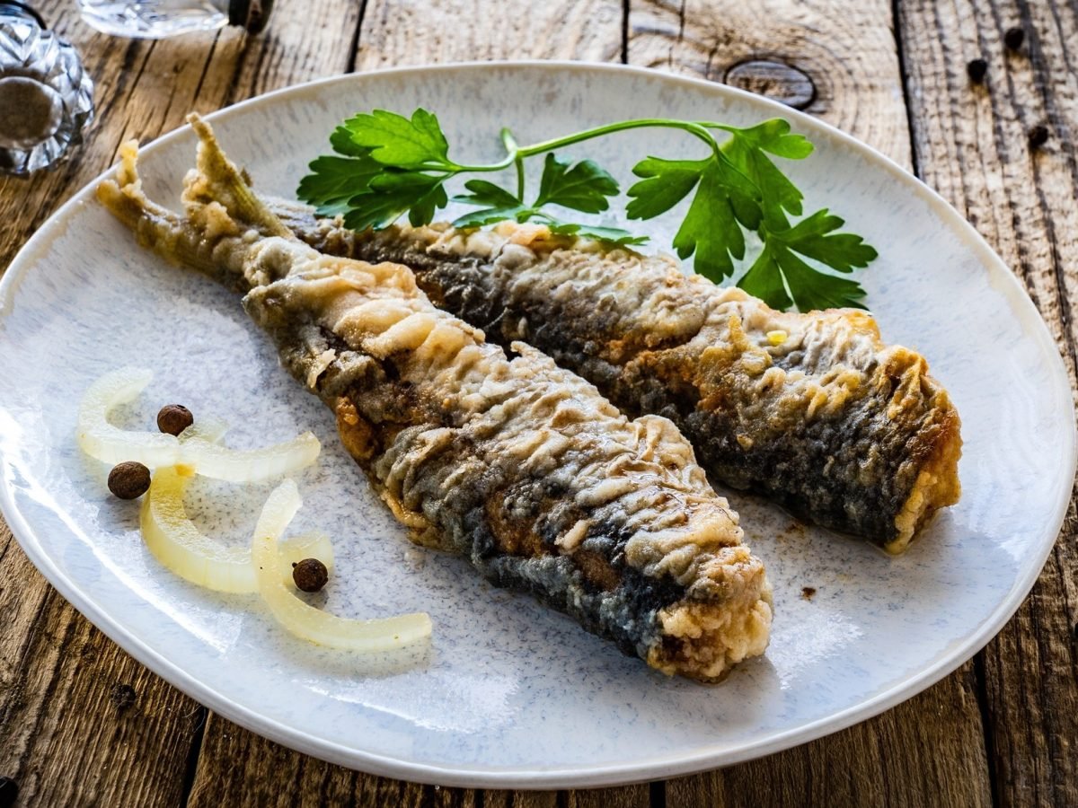 Draufsicht schräg: Ein weißer Teller mit zwei Bratheringen liegt auf einem rustikalen Untergrund aus Holz. neben den Heringsfilets liegen rohe Zwiebelringe und frische Petersilie.