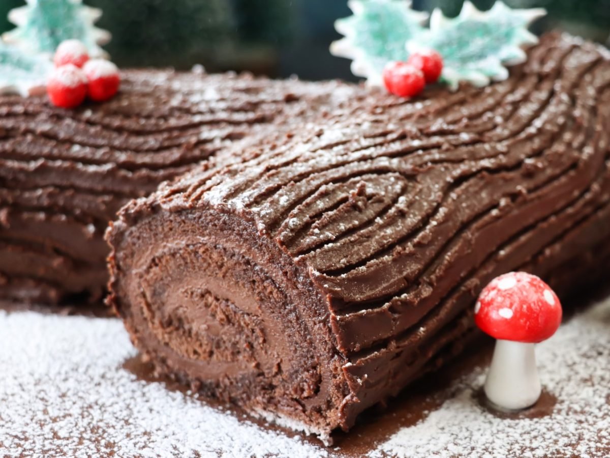 Bûche de Noel ohne Mehl auf Puderzucker, rechts davon Zuckerfiguren.