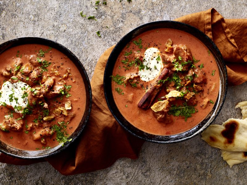 Zwei Schüsseln Butter-Chicken-Soup, daneben Stücke Fladenbrot auf grauem Untergrund.