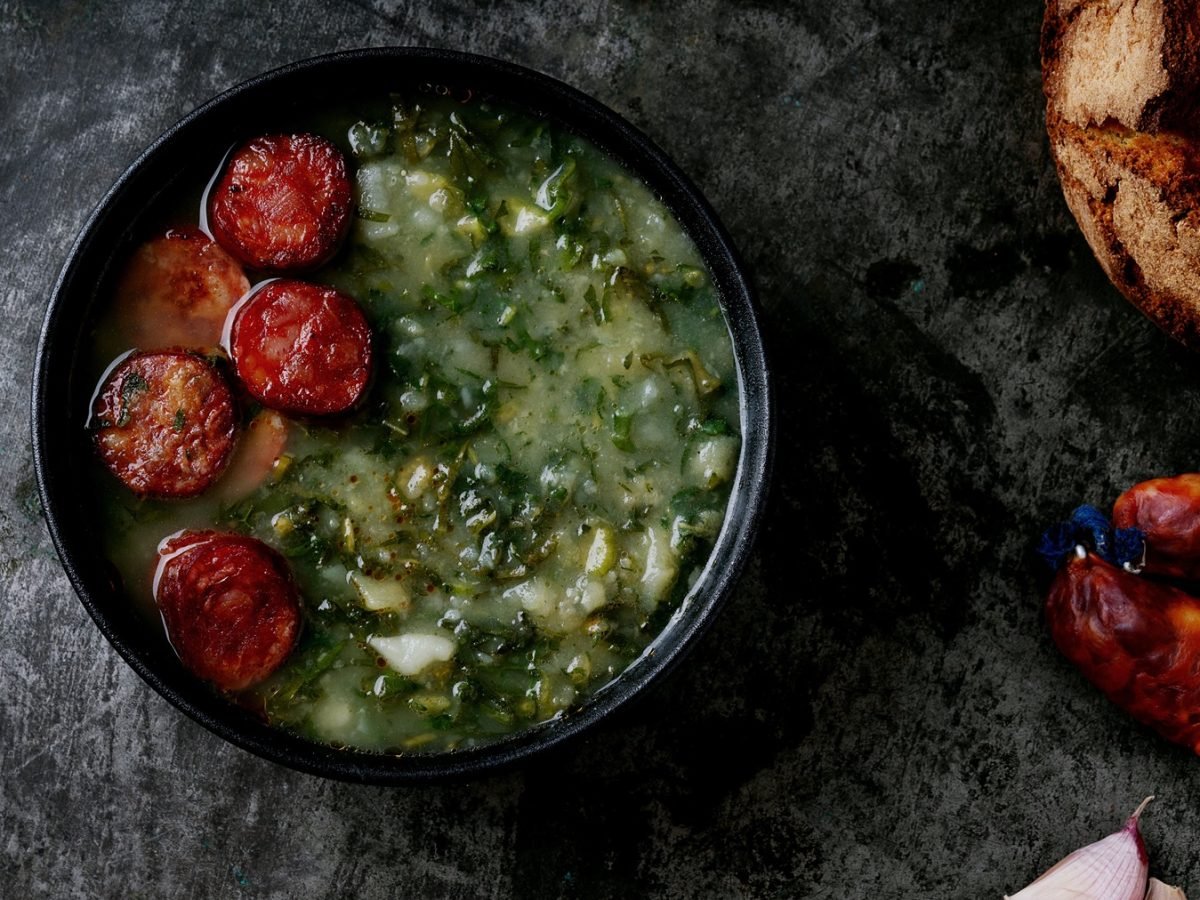 Eine schwarze Schale mit der Suppe auf einem grauen Steintisch, mit Knoblauchzehen, Chorizo und Sauerteigbrot im rechten Bildrand. Alles direkt von oben fotografiert.