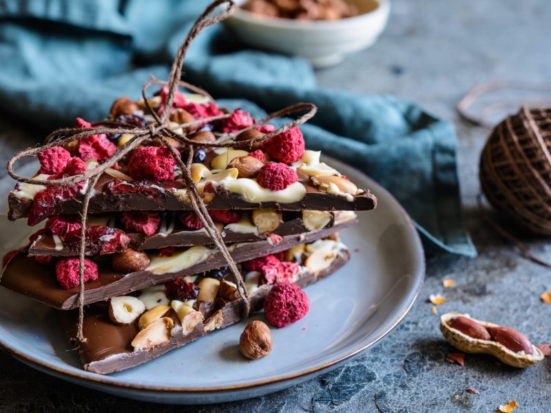 Frontalsicht auf mehrere Stücke Chocolate Bark mit Trockenfrüchten und Nüsse, die zu einem Paket geschnürt sind. Daneben eine Spule mit Paketband.