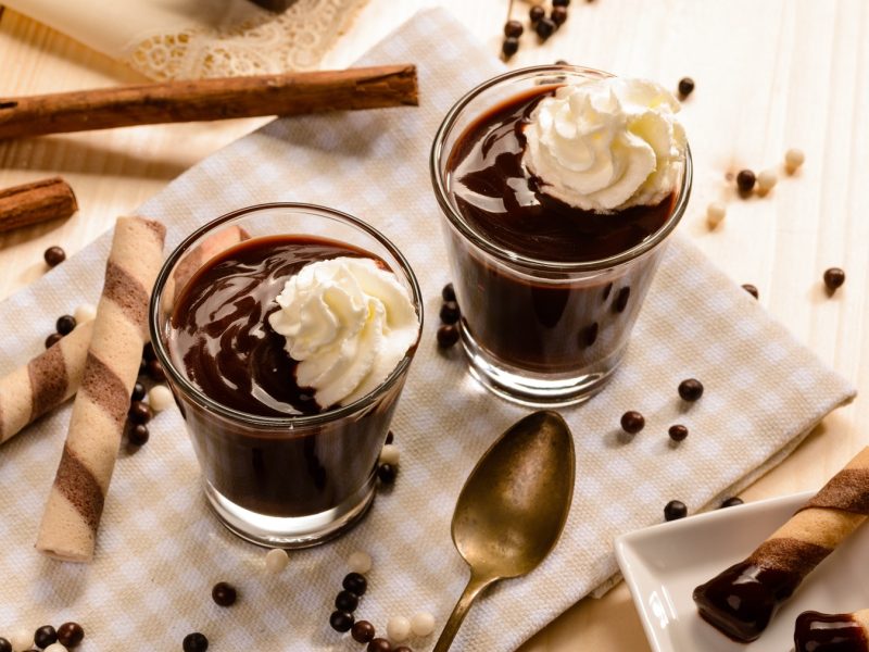 Draufsicht auf zwei Gläser mit cioccolata calda, mit Schlagsahnehäubbchen, daneben Schokoperlen und Plätzchen.