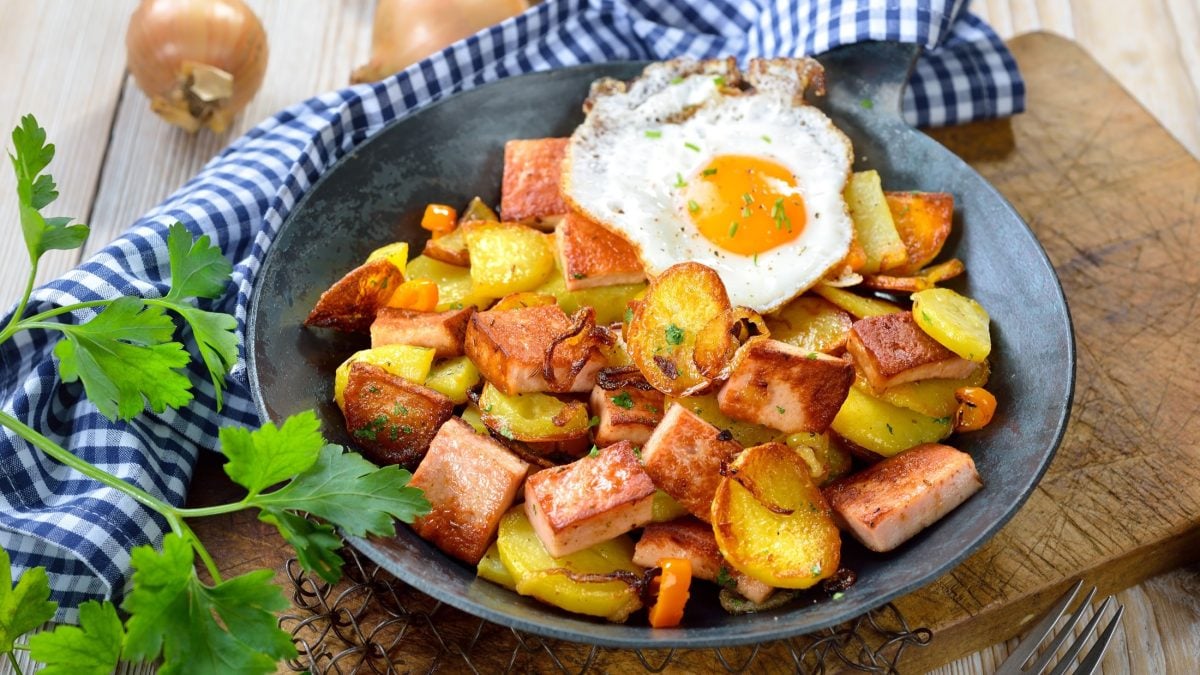 Leberkäse mit Spiegelei und Bratkartoffeln in einem großem Teller. Der steht auf einem Untergrund aus rustikalem Holz. / Draufsicht,