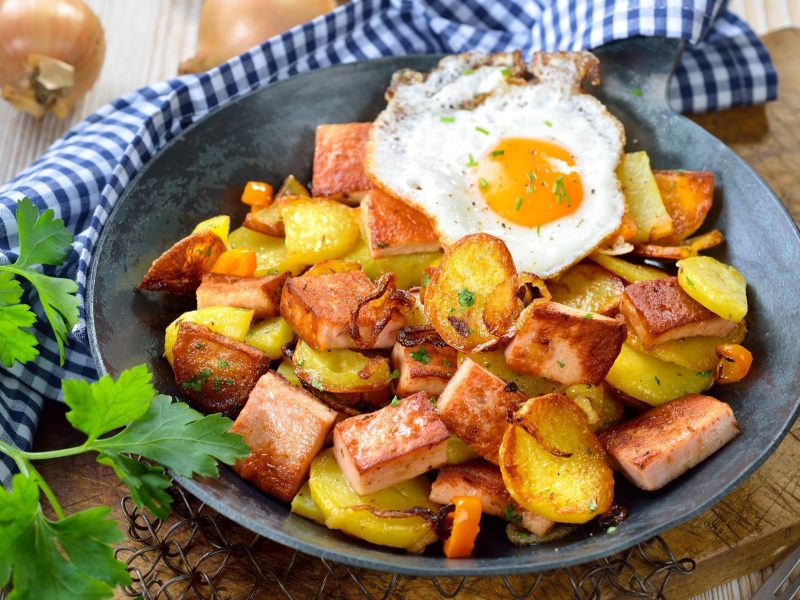 Leberkäse mit Spiegelei und Bratkartoffeln in einem großem Teller. Der steht auf einem Untergrund aus rustikalem Holz. / Draufsicht,