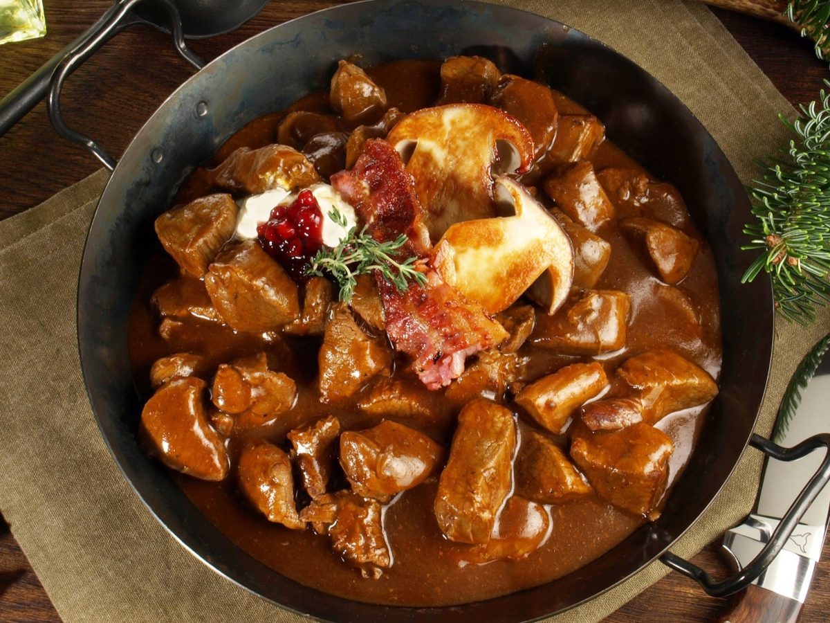 Draufsicht: Ein gusseiserner Topf in dem ein Rehragout ist, steht auf einem groben Leinentuch. Das Liegt auf einem Holztisch. Drum herum liegen weihnachtlich anmutende Tannenzweige, Hagebutten, ein aufgeklapptes Taschenmesser.