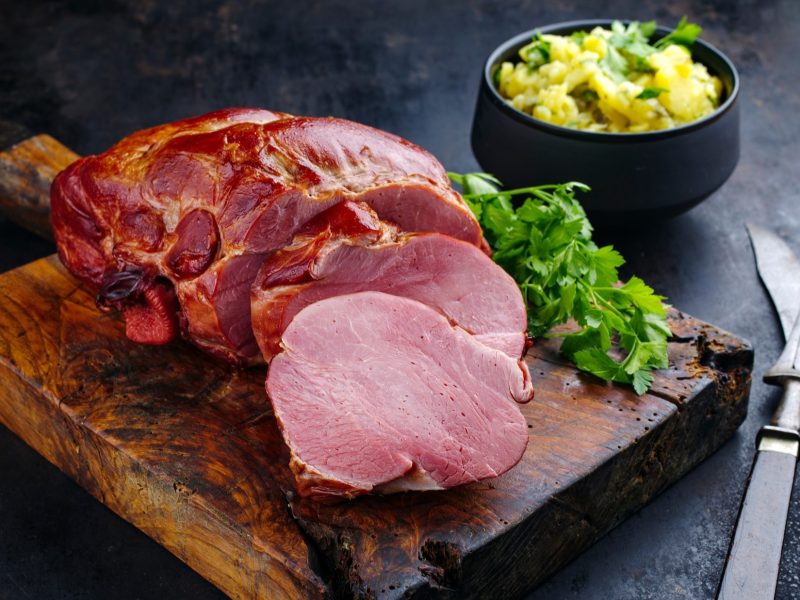 Schäufele, angeschnitten, auf einem rustikalem Brettchen. Dabei handelt es sich um ein großes Stück Schweinefleisch, dass im Wasser gegart wurde. Rechts davon liegt ein Strauß frische Petersilie. Das Holzbrettchen liegt auf einem schwarzen Untergrund. Daneben ist eine Schale mit Kartoffelsalat und ein Besteck.
