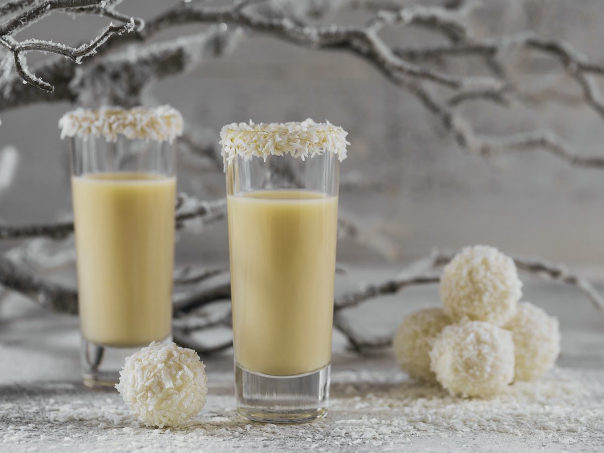 Zwei Shotgläser mit Eierlikör und darunter die Eierlikörpralinen. Alles auf einem winterlich dekorierten Tisch mit Kunstschnee und Ästen.