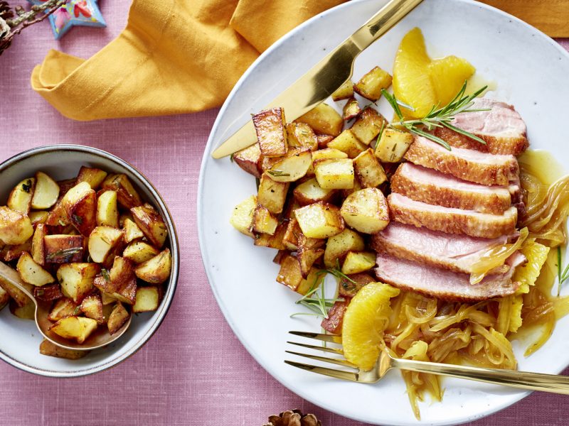 Draufsicht: Entenbrust mit Orangensauce und Bratkartoffeln auf einem Teller, daneben Schüssel mit mehr Bratkartoffeln, rosa Untergrund.