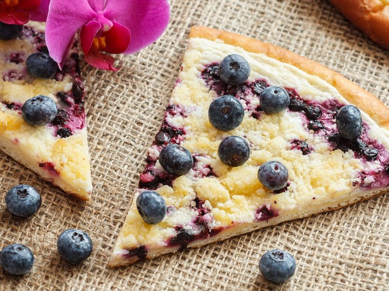 Stück Frgál mit Blaubeeren und Streusel auf einem Sisal-Untergrund, im Hintergrund eine pinke Orchidee.