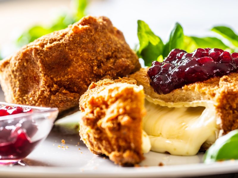 Nahaufnahme vom angeschnittenen Back-Camembert, mit Preiselbeeren garniert. Alles von der Seite und vor hellem Hintergrund fotografiert. Links daneben noch ein kleines Schälchen mit Preiselbeeren und unscharf ein paar grüne Kräuter im Hintergrund.