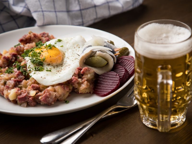 Draufsicht, schräg: Ein Teller mit Hamburger Labskaus steht auf einem Holztisch. Daneben liegt ein Besteck. Neben den steht ein Glas Bier.