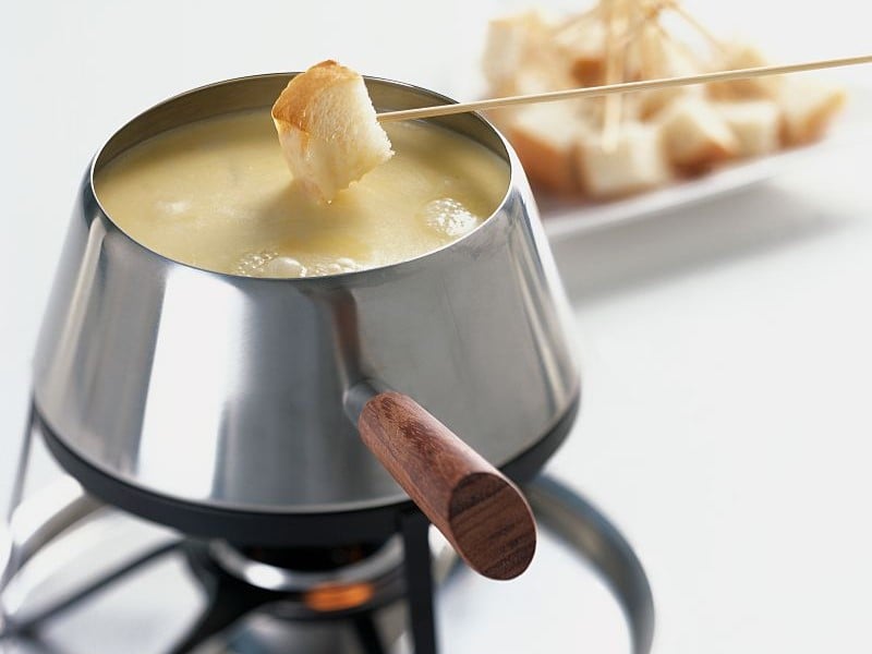 Ein silberner Topf mit Käsefondue auf einem hellen Tisch mit mehr Spießen im Hintergrund.