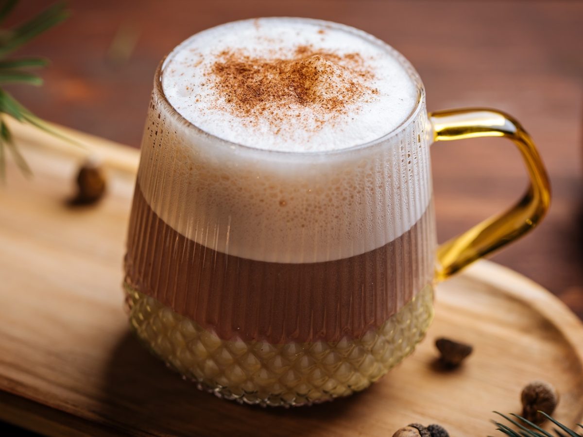 Ein Glas mit dem Eierlikör-Macchiato auf einem hellen Holzbrett von schräg oben fotografiert. Im Hintergrund winterliche Dekoration.