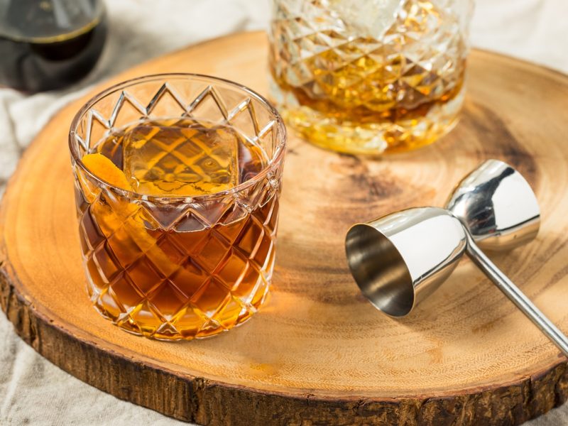 Zwei Gläser mit Maple Oldfashioned auf einer Holzscheibe, daneben Barzubehör.