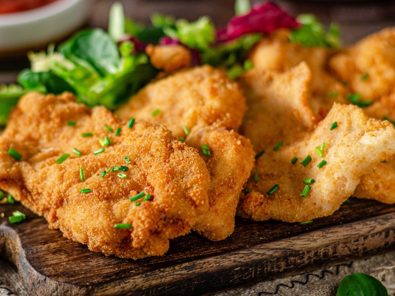 Das Münchner Schnitzel auf einem dunklen Holzbrett und dunklem Holztisch. Alles von der Seite fotografiert mit Salat und Co im Hintergrund.