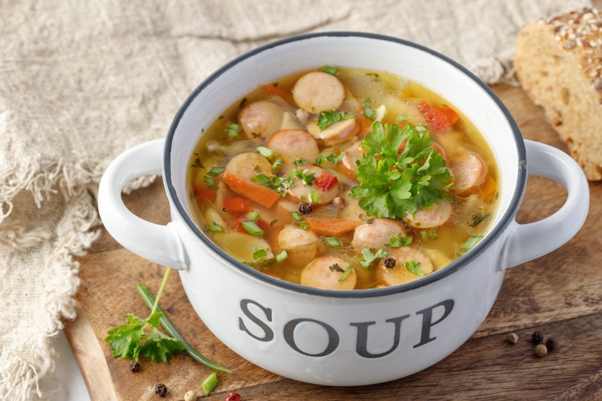 Nudelsuppe mit Würstchen in einer weißen Schüssel auf einem hölzernem Brett.