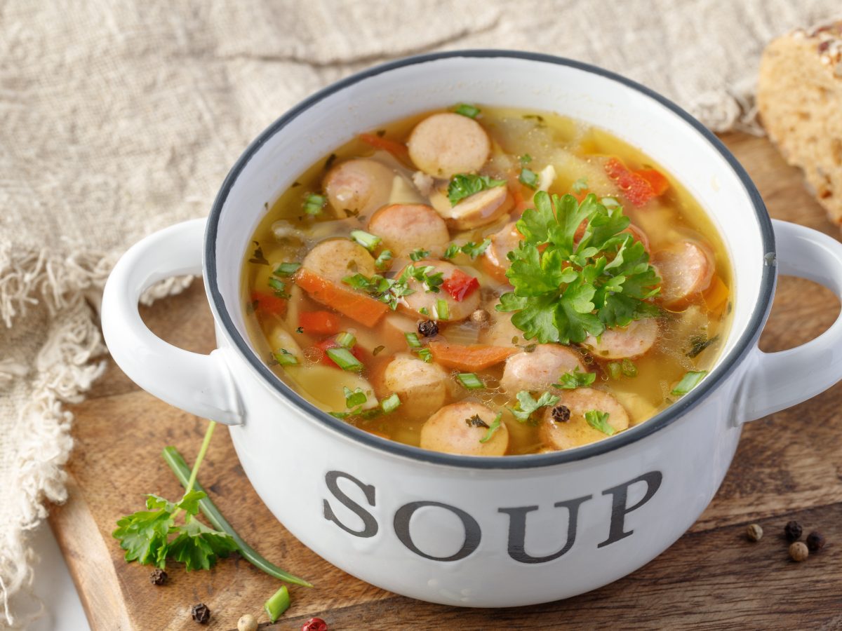 Nudelsuppe mit Würstchen in einer weißen Schüssel auf einem hölzernem Brett.