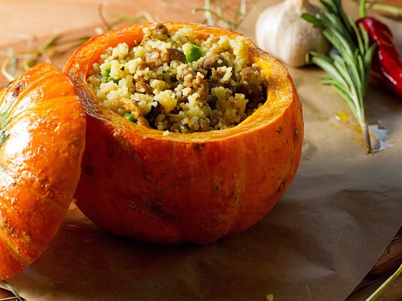 Ein Holztisch mit dem Kürbis auf Backpapier rund vielen Kräutern wie Rosmarin, Chili und Knoblauch. Alles von der Seite fotografiert.