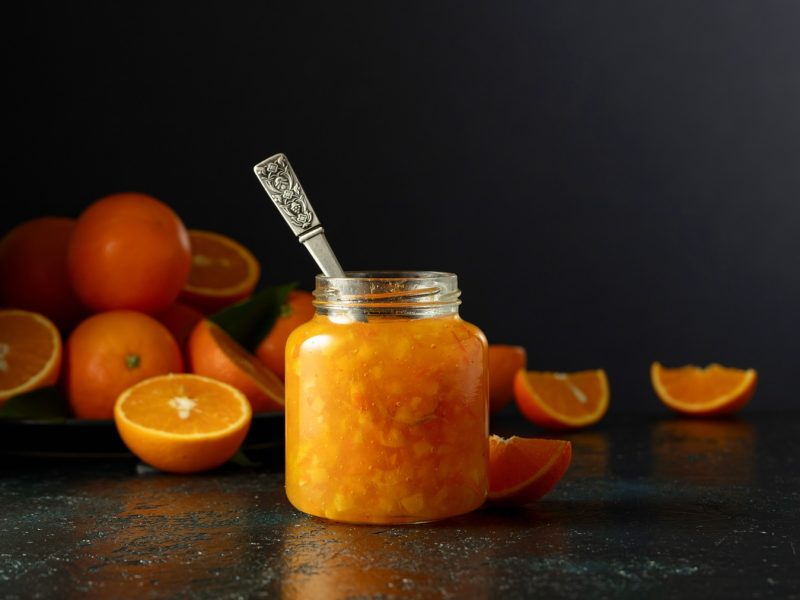 Ein Glas mit der Orangenmarmelade und ein paar halbierten Orangen auf dunklem Untergrund und vor schwarzem Hintergrund.