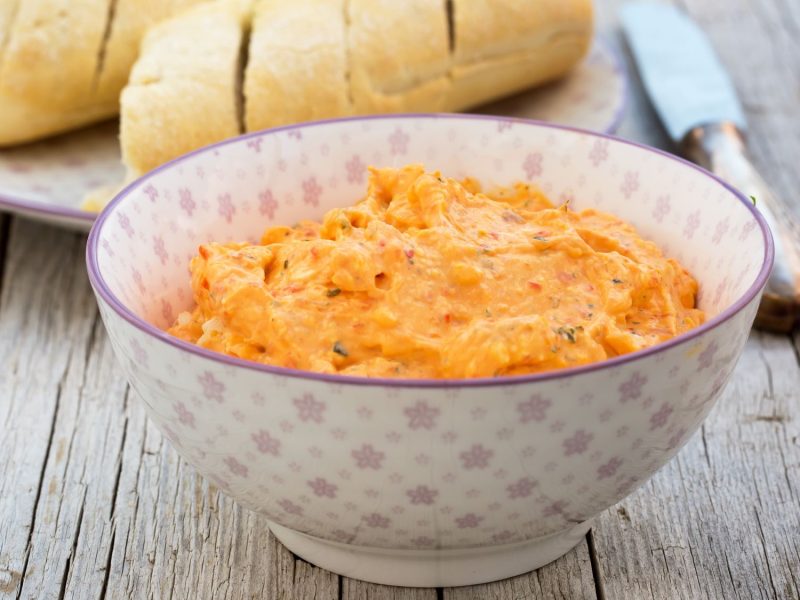 Selbstgemachter Paprika-Feta-Dip mit Kräutern in dekorativer Schale, serviert auf einer Holzoberfläche mit Brotscheiben.