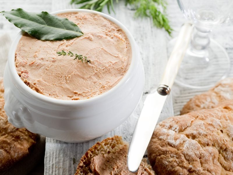 Draufsicht auf ein weißes Porzellangefäß mit Pâté di fegato, darauf ein Lorbeerblatt. Daneben frischen Brot und ein Messer.