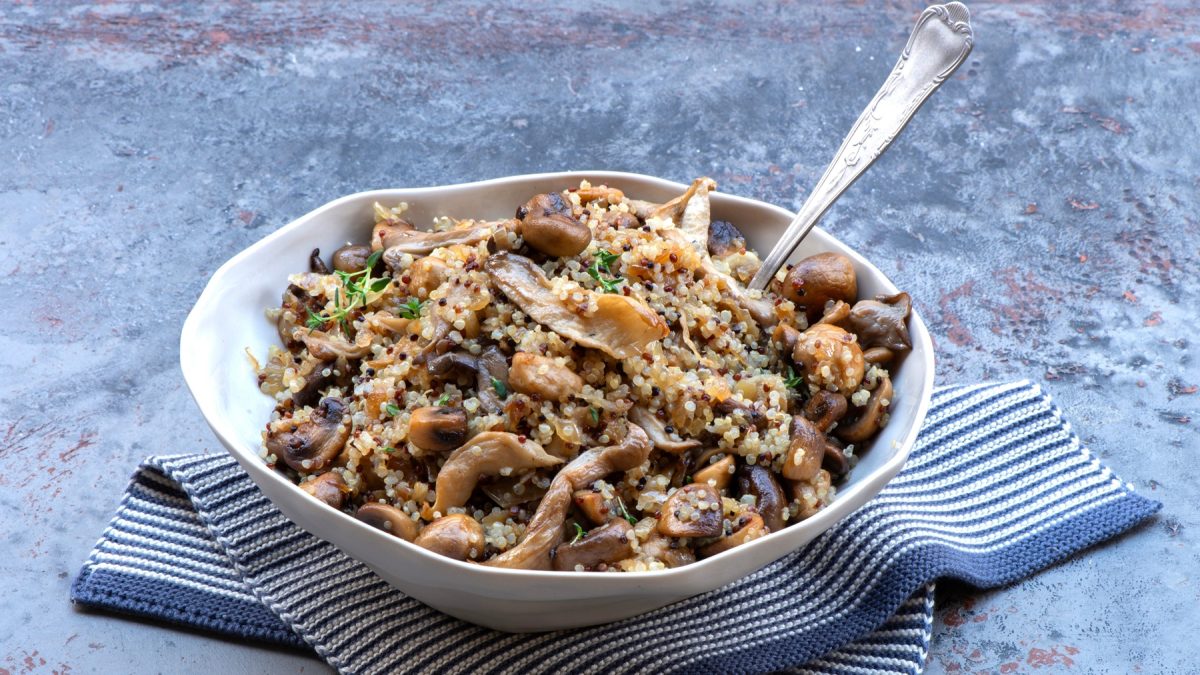 Ein weißer Teller mit der Pilz-Quinoa-Pfanne auf einem blauen Marmortisch mit weiß-blauem Tuch. Alles von der Seite fotografiert.