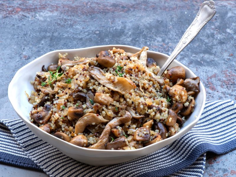 Ein weißer Teller mit der Pilz-Quinoa-Pfanne auf einem blauen Marmortisch mit weiß-blauem Tuch. Alles von der Seite fotografiert.