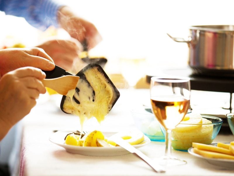 Eine Hand schabt geschmolzenen Käse aus einer Raclette-Pfanne auf einen Teller mit Kartoffeln, daneben ein Glas Weißwein.