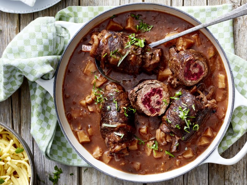 Draufsicht: Rinderroulade mit Rote-Bete-Füllung in einem Topf mit brauner Sauce, daneben Spätzle mit hellem, rustikalen Hintergrund.