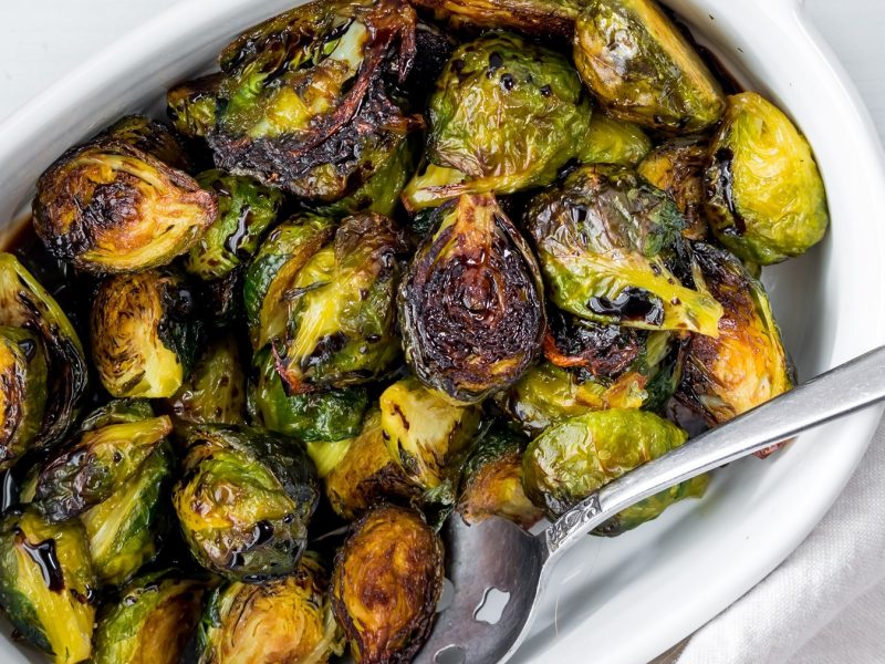 Eine weiße Auflaufform mit dem Rosenkohl aus dem Airfryer und einem silbernen Löffel auf einem weißen Tisch. Alles sehr nah und direkt von oben fotografiert.