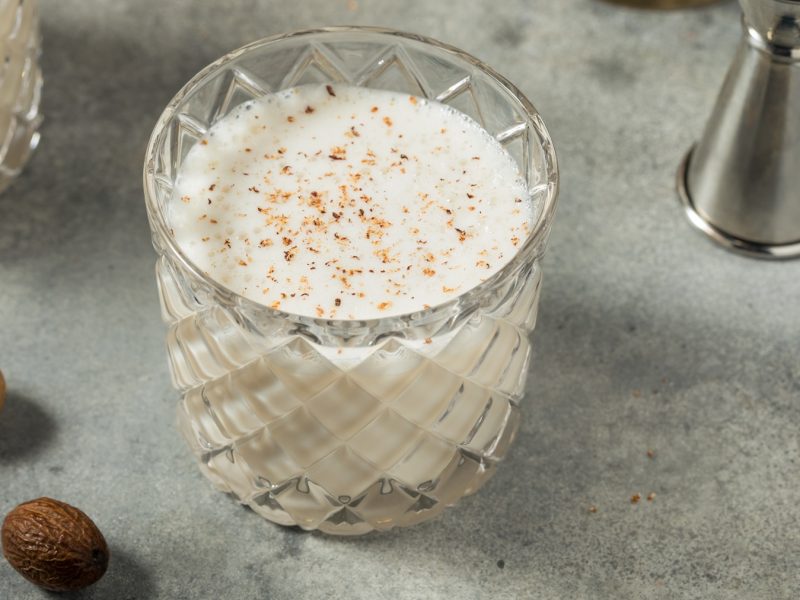 Glas mit Rum Milk Punch vor grauem Hintergrund, daneben frische Muskatnüsse.