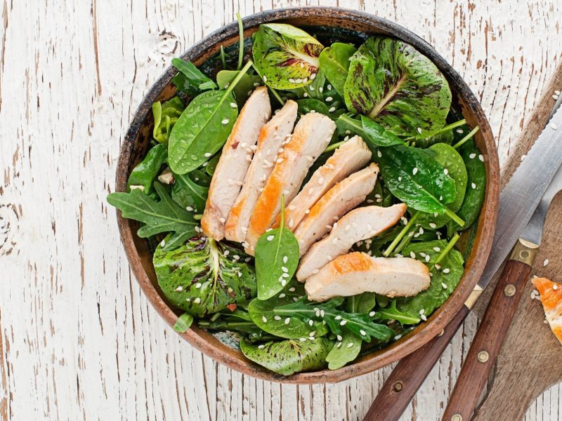 Eine Schüssel mit einem Salat mit Hähnchenbrust und Mandel-Dressing steht auf einem hellem Untergrund aus Holz. Daneben liegt ein Holzbrettchen mit Besteck. / Draufblick