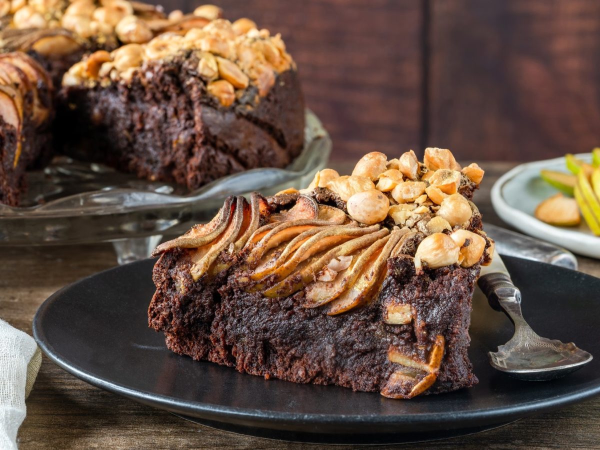Frontal: eine angeschnittene Schokoladen-Torte mit Nüssen und Äpfeln. Sowie ein Teller mit einem Stück Torte. Im Hintergrund sind geschnittene Äpfel und ein Glas Tee.