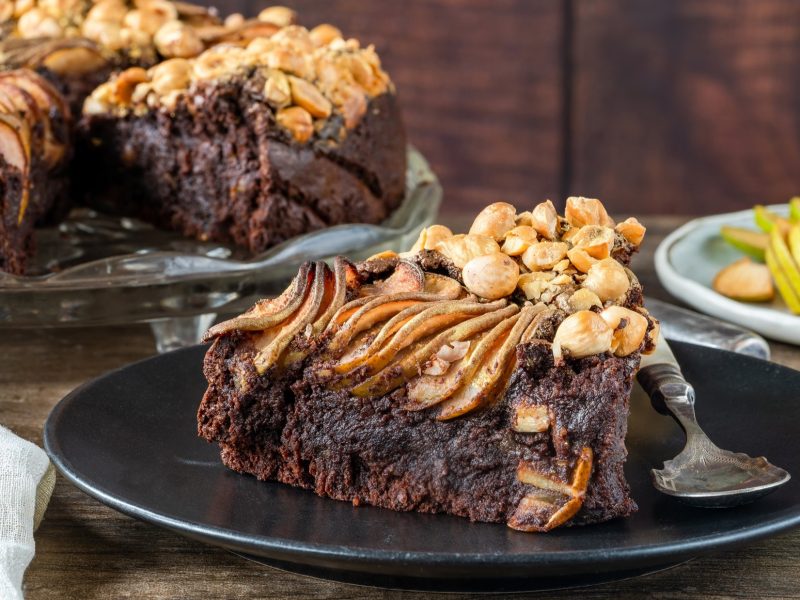 Frontal: eine angeschnittene Schokoladen-Torte mit Nüssen und Äpfeln. Sowie ein Teller mit einem Stück Torte. Im Hintergrund sind geschnittene Äpfel und ein Glas Tee.