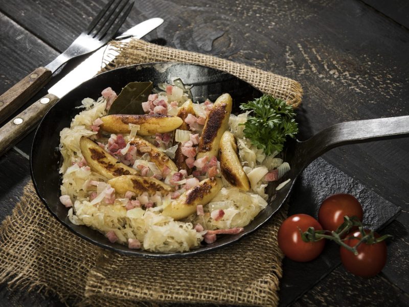 Schupfnudeln mit Sauerkraut in einer rustikalen Pfanne, die auf einem rustikalen Tisch aus Holz steht. Neben der Pfanne liegen Besteck sowie frische Tomaten.