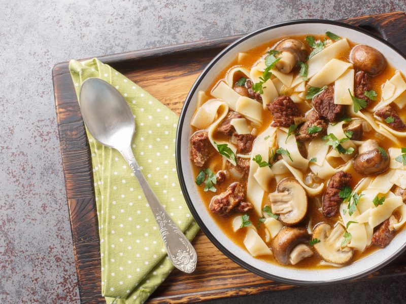 Weißer Teller gefüllt mit einer Stroganoff-Nudelsuppe. Dieser steht auf einem Holzbrett, auf dem auch ein Löffel und eine grüne Serviette liegt.