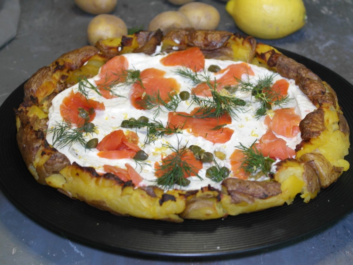 Ein schwarzer Teller, mit der Kartoffelpizza belegt mit Lachs, Kapern, Dill und Zitronenabrieb. Alles von leicht schräg oben auf grauem Steinuntergrund fotografiert.