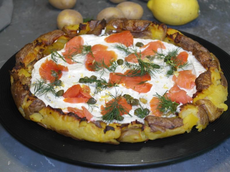 Ein schwarzer Teller, mit der Kartoffelpizza belegt mit Lachs, Kapern, Dill und Zitronenabrieb. Alles von leicht schräg oben auf grauem Steinuntergrund fotografiert.