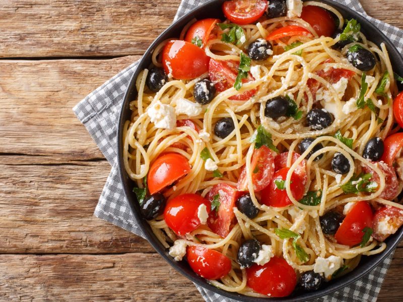 Pasta al greco mit schwarzen Oliven, Kirschtomaten, Fetakäse und frischen Kräutern in einer rustikalen Schale.
