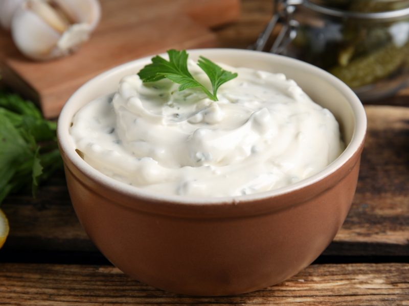 Ein braunes Schälchen mit der veganen Remoulade auf einem Holztisch, mit enthaltenen Zutaten wie Kapern, Dill, Zitrone, Knoblauch und Cornichons. Alles von der Seite fotografiert.