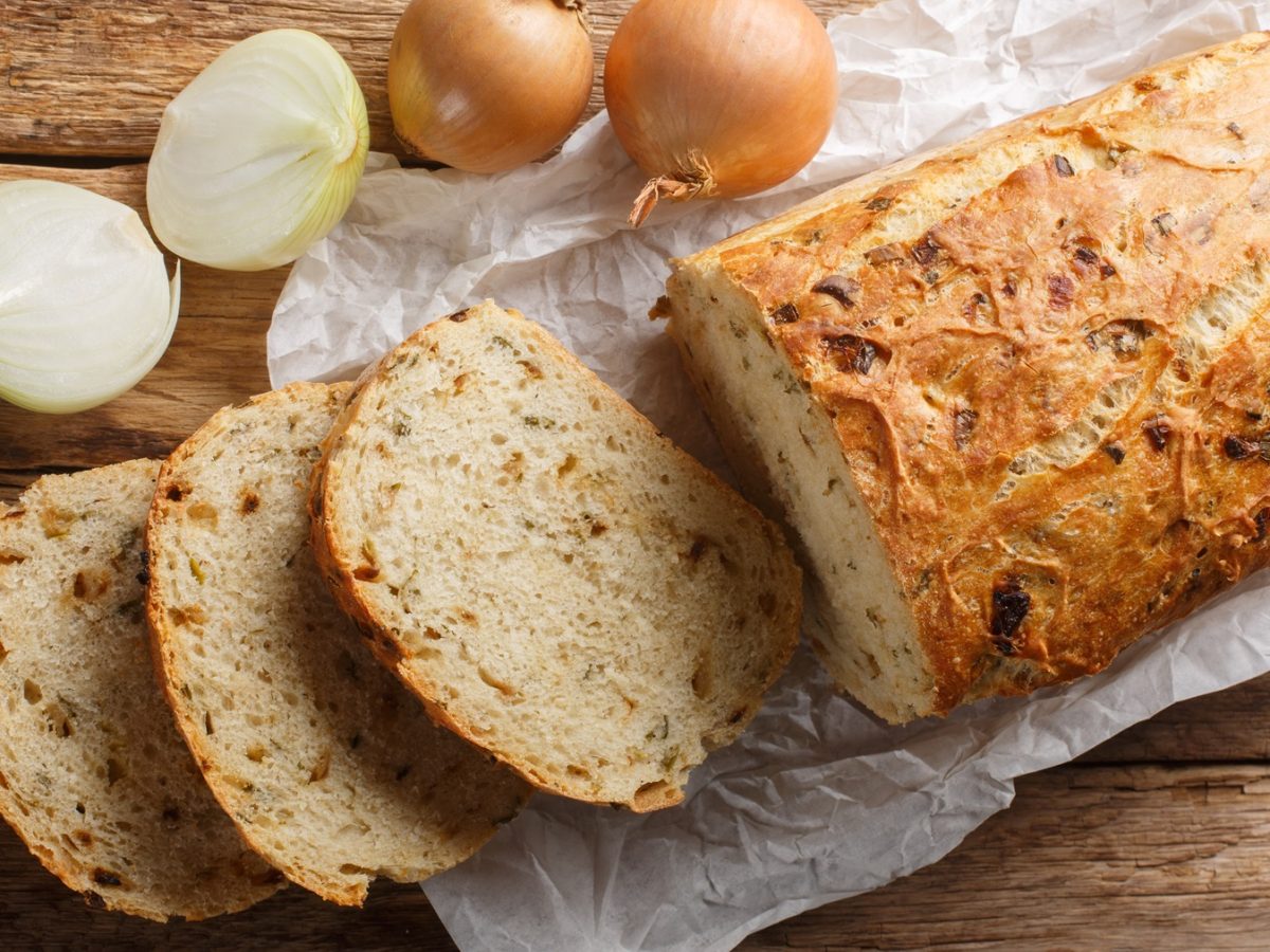 Draufsicht auf ein angeschnittenes Zwiebelbrot, daneben eine halbierte und eine ganze frische Zwiebel.